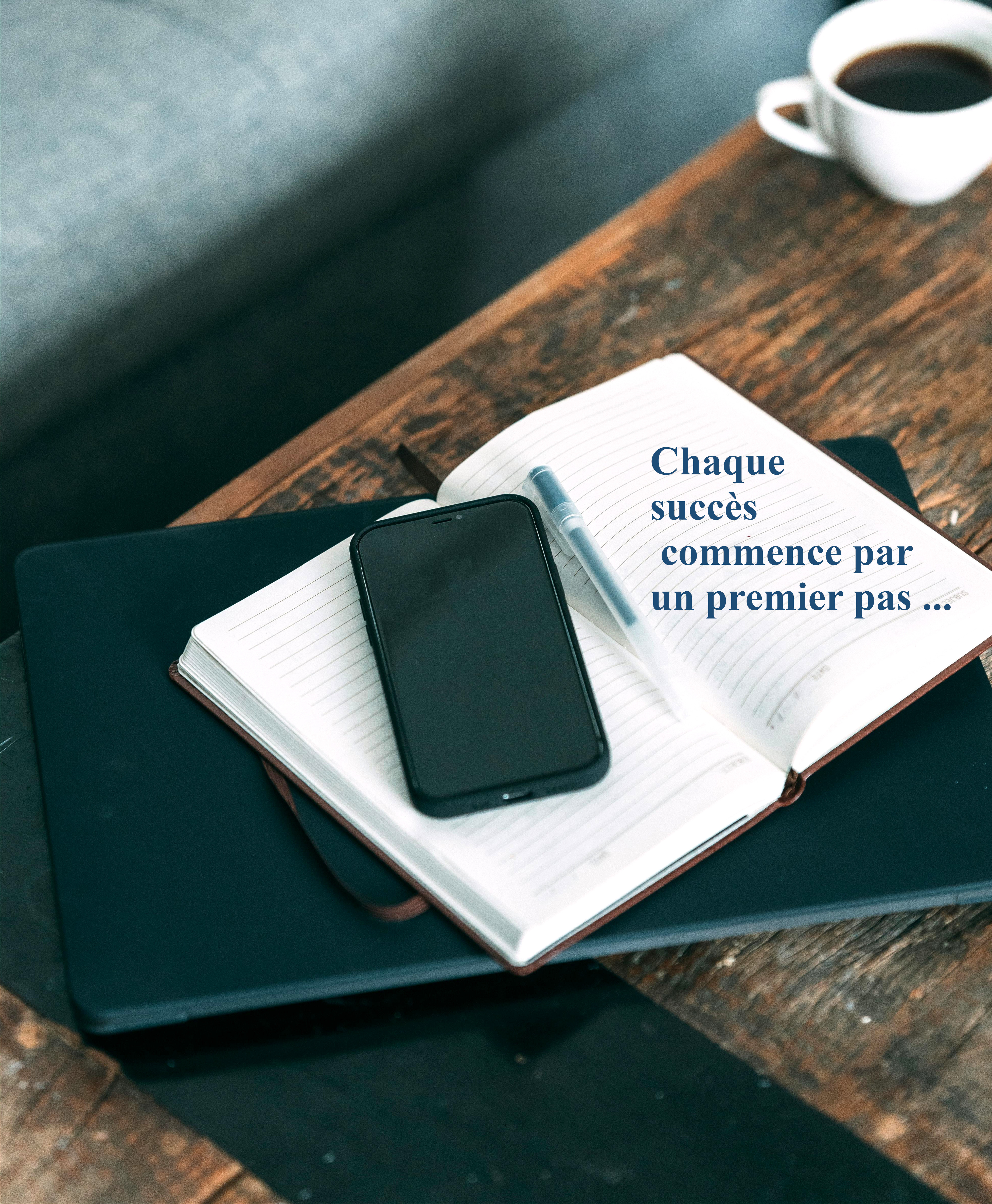 Photo d'outils permettant de prendre contacte: téléphone, carnet de notes sur lequel il est inscrit "Chaque succès commence par un premier pas ...", et ordinateur portable, sur un bureau avec un café. 